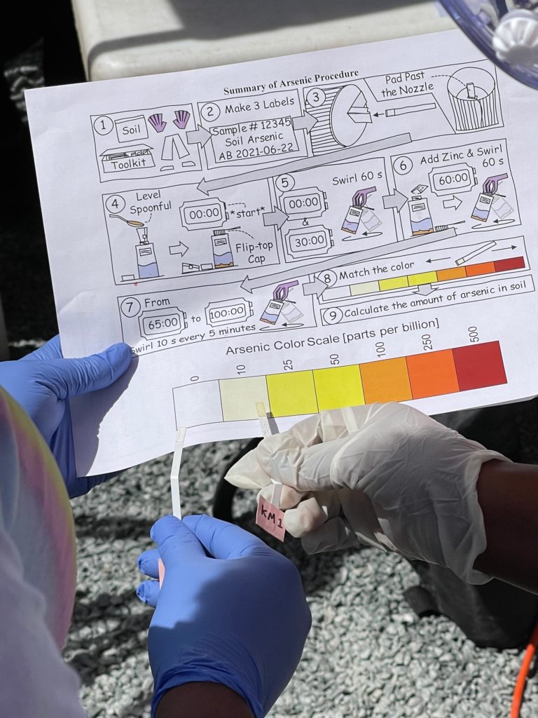 Two people compare a test strip to a color scale to evaluate arsenic concentration.